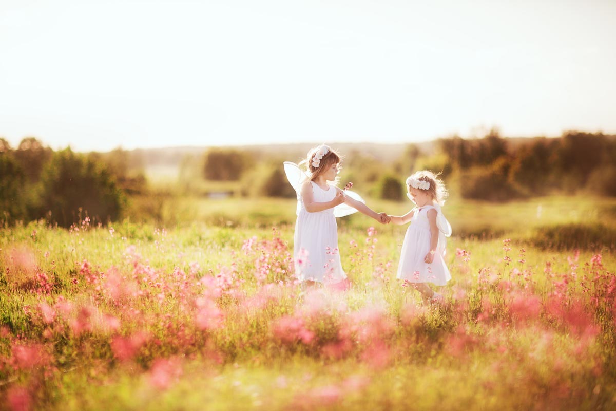 A white 5 year old and her younger sister in white dresses and flower crowns hold hands in a field of flowers on a sunny day